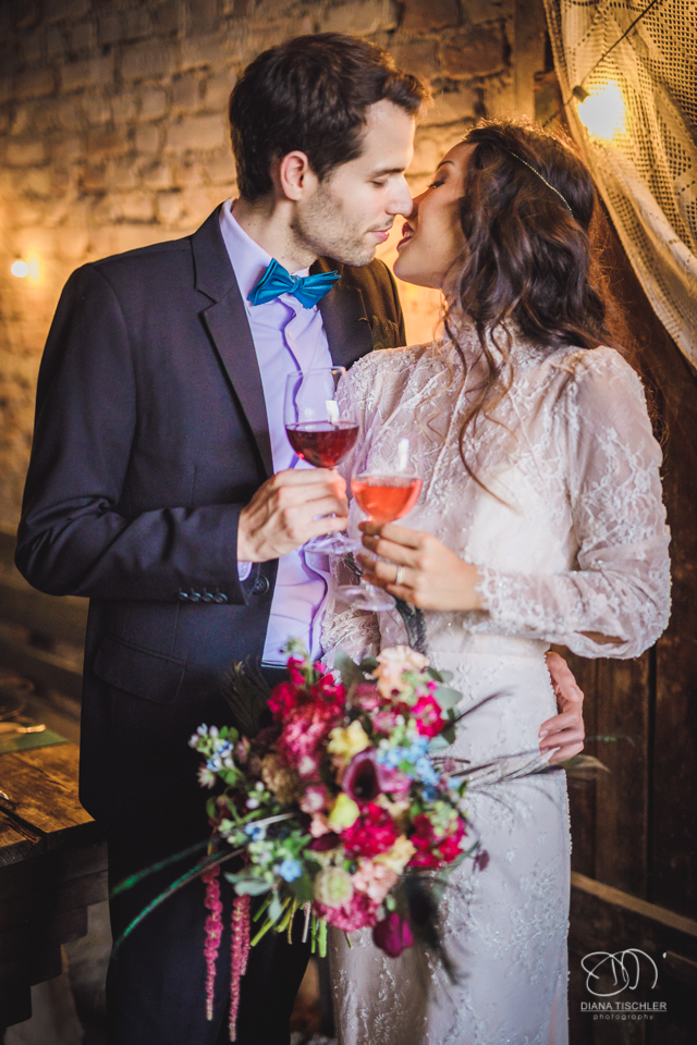 Brautpaar mit tollem buntem Brautstrauss stoesst mit Wein an bei einer Hochzeit in einer Scheune