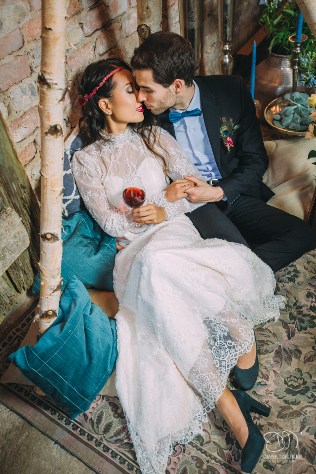 Brautpaar in Kissen und Decken stoesst mit Wein an sich bei einer Hochzeit in einer Scheune