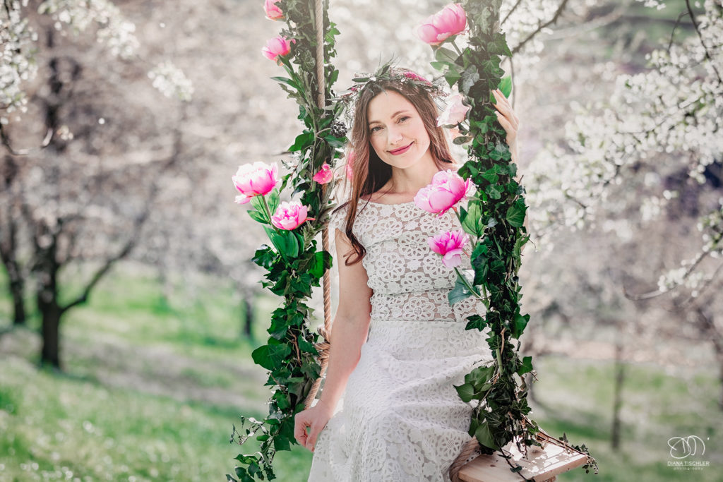 Braut auf Schaukel mit Blumen unter Kirschbaum auf Wiese