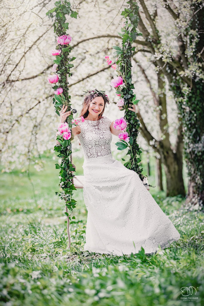 Braut auf Schaukel mit Blumen unter Kirschbaum auf Wiese